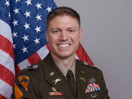 Person wearing military uniform, smiling in front of American flag
