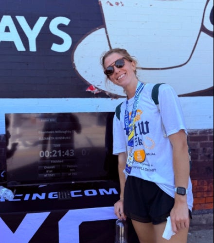 Elizabeth Ginsburg in sunglasses and wearing a race medal stands in front of a computer screen displaying her race time.