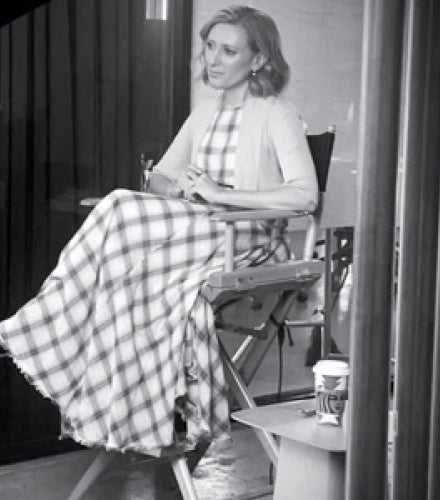 Black and white image of a woman seated in a director's chair, wearing a long checkered dress. She appears calm and focused, with a coffee cup on a side table.