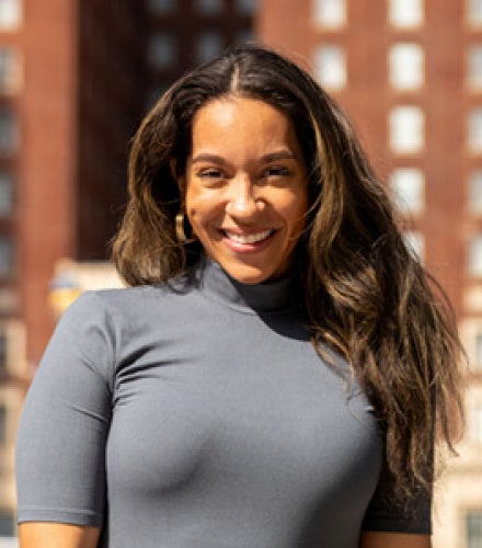 Smiling woman in a gray dress stands outside with tall red brick building in the background. Bright sunlight creates a warm and cheerful atmosphere.