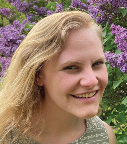 A smiling woman with blonde hair stands in front of blooming lilac bushes. She wears a patterned green dress, exuding a joyful and serene vibe.