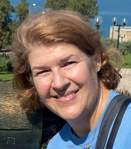 A smiling woman with short brown hair wearing a blue shirt and necklace, outdoors on a sunny day with trees and lampposts in the background.