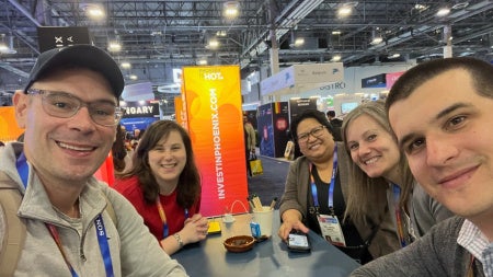 A group of people sit around a table at a busy expo. The setting features bright signage and various booths and banners in the background. 