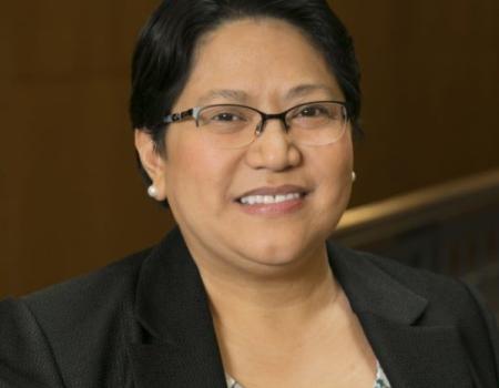 Anna Samia is wearing a black blazer and a patterned blouse, and sitting in front of a wood-paneled background.