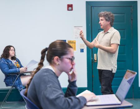 A classroom setting with a teacher standing and speaking. Two students, one focused on papers and the other on a laptop, listen attentively.