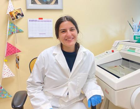 A woman wearing a lab coat and blue gloves smiling in a lab. She is next to scientific equipment.