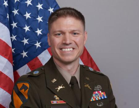 Person wearing military uniform, smiling in front of American flag