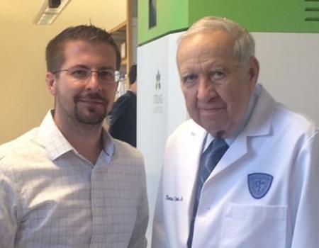 Two men standing side by side, one on left wearing dress shirt and on right white lab coat