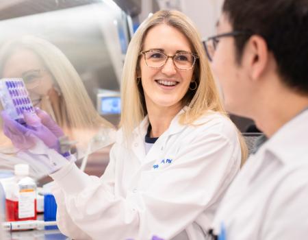 A woman in a lab coat and gloves smiles while holding a test tube rack, engaging with a colleague.
