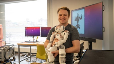 Austin holds a robot in front of a large wall-mounted screen displaying a robot silhouette
