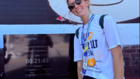 Elizabeth Ginsburg in sunglasses and wearing a race medal stands in front of a computer screen displaying her race time.
