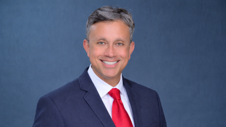 Andres Pinto poses in a blue jacket and red tie in front of a blue background.