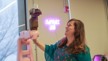 A scientist smiles and looks at a pink robotic arm with a sock puppet