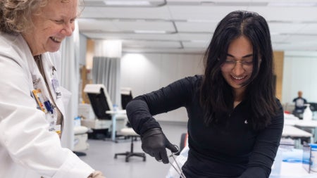 A student in a black shirt practice sutures while a professor in a white lab coat watches and smiles.