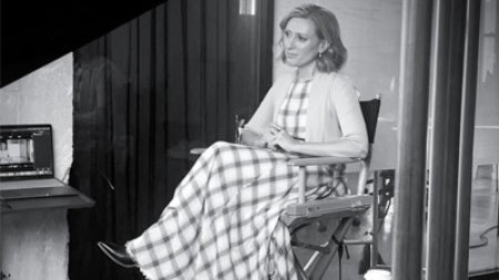 Black and white image of a woman seated in a director's chair, wearing a long checkered dress. She appears calm and focused, with a coffee cup on a side table.