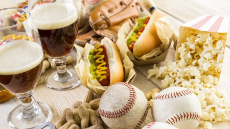 Spread featuring hot dogs, popcorn, peanuts, two glasses of beer, baseballs, a glove, and a bottle opener on a wooden surface.