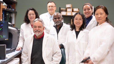 Seven researchers in white lab coats pose together inside a biomedical laboratory. Shelves lined with binders and lab supplies surround the group, while a computer monitor on the left displays colorful microscopic cell images. 