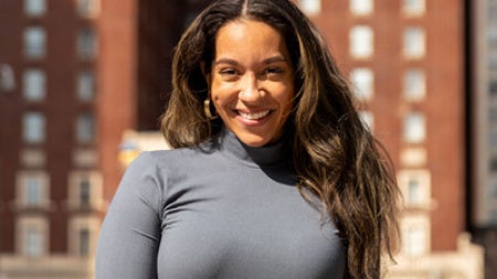 Smiling woman in a gray dress stands outside with tall red brick building in the background. Bright sunlight creates a warm and cheerful atmosphere.