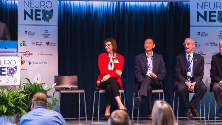 Panel of five professionals seated on stage at NeuroNEO 2026 conference. The speaker stands at a podium with banners and plants nearby.