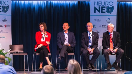 Panel of five professionals seated on stage at NeuroNEO 2026 conference. The speaker stands at a podium with banners and plants nearby.
