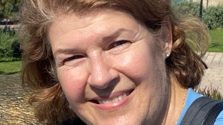 A smiling woman with short brown hair wearing a blue shirt and necklace, outdoors on a sunny day with trees and lampposts in the background.