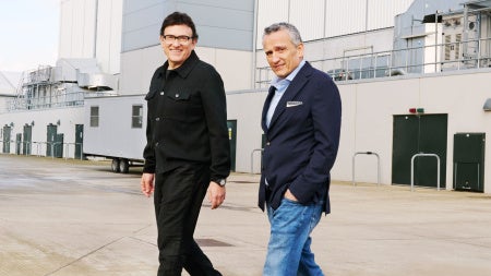 Two men are walking on a concrete surface outside an industrial building. One wears a black jacket, the other a navy blazer and jeans. Both are smiling.