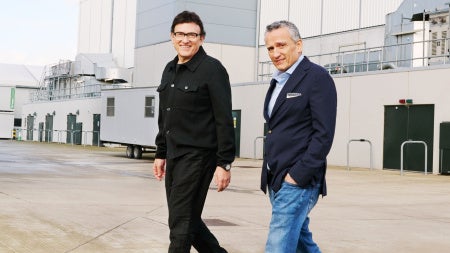 Two men are walking on a concrete surface outside an industrial building. One wears a black jacket, the other a navy blazer and jeans. Both are smiling.