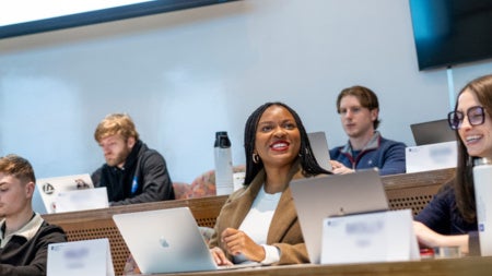 Seven students sit in a tiered classroom in the Peter B. Lewis Building. Two students smile and interact with each other while the others focus on their laptops. 