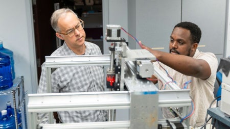 Two men, focused and engaged, work together on a complex machine in a lab. One points attentively while the other observes, discussing adjustments.
