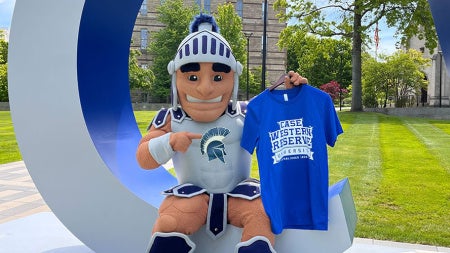 Photo of Spartie sitting in the C of the big CWRU letters on campus while holding up a Blue CWRU shirt