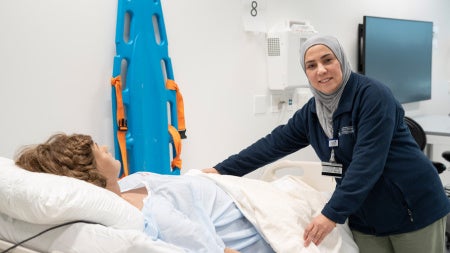 A nursing professor is placing a blanket on a pregnant, high-fidelity mannequin lying on a bed. The background mimics the hospital setting.  