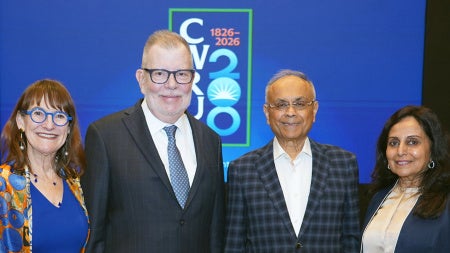 Four people stand smiling against a blue backdrop with "CV 20-26" text. They are dressed in formal attire, reflecting a professional event.