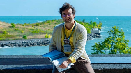 Photo of Sebastian Correa holding a book while posing with a tropical area behind him
