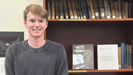 Aidan Selkirk poses for a photo with a copy of his artwork created with an aerosol jet printer framed on a shelf next to him