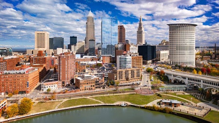 Photo of the Cleveland skyline with clouds set against a bright blue sky and the Cuyahoga River in the foreground