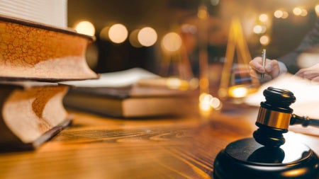 A gavel and books sit on a desk in front of a man reading a book.