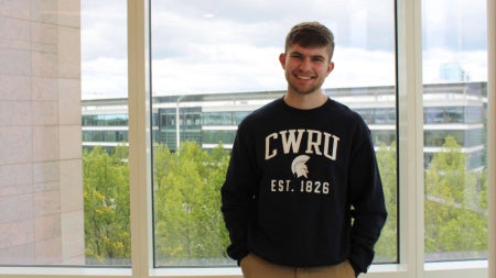 Jacob Hoffa stands in front of a window in front of Samson Pavilion wearing a CWRU sweatshirt.