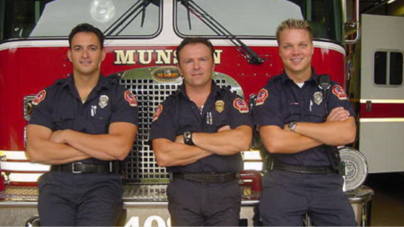 Three men stand in front of a fire truck with arms crossed.