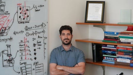 A researcher stands beside a whiteboard covered with diagrams, equations, and notes about imaging, scattering, and polymers. On his right, there are shelves filled with scientific textbooks and framed research awards.