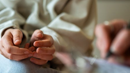Photo showing the closeup of a teenager with her hands in her lap as a person next to her takes notes with a pen and notepad