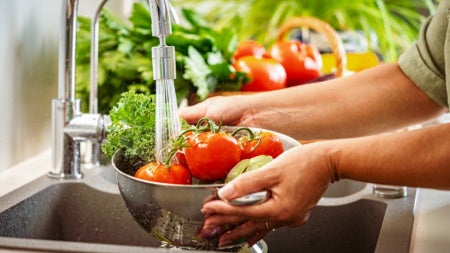 Hands holding a colander full of fresh vegetables under running water in kitchen sink