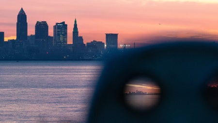 Photo taken from behind a photo viewfinder at a park showing the Cleveland skyline and Lake Erie set against a pinkish blue sky, with one eye hole focused on the view