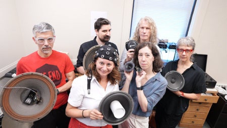 Six experts in the field of music, cognitive sciences, and audio engineering pose in a lab space while holding speakers and other audio equipment, including one person wearing an EEG cap with sensors.