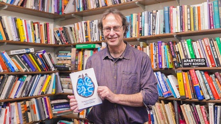 The professor stands in front of shelves filled with books, holding a copy of his book: The Empire of Depression.