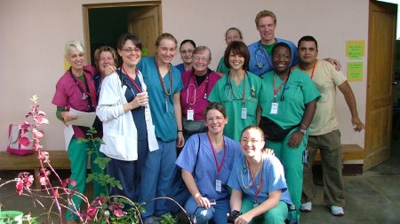 A smiling, diverse healthcare team in scrubs and white coats poses in a hospital doorway beside plants.