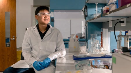 A smiling Joshua Kuwana in a white lab coat and blue gloves sits beside glassware and equipment in a bright laboratory.