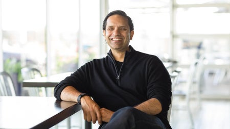 Randhir-Sethi in a black sweater sits casually on a wooden railing in a bright, modern office, conveying a warm, relaxed confidence.