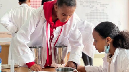 Two students in white lab coats collaborate at a table of scientific equipment, focused on an experiment in a bright classroom with a world map.