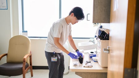 Andrew Liang, wearing a white shirt and blue gloves, prepares equipment at a counter in a bright clinic room, focused and calm.