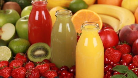 Three bottles of red, green and yellow fruit juice bottles surrounded on a table by fresh fruits